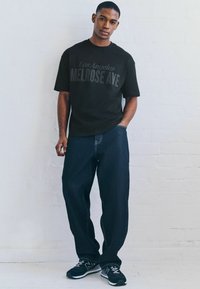 Crna majica s natpisom "Los Angeles MELROSE AVE" ispisana tamnosivom bojom. Nošena uz tamnoplave široke traperice i crne sportske cipele.