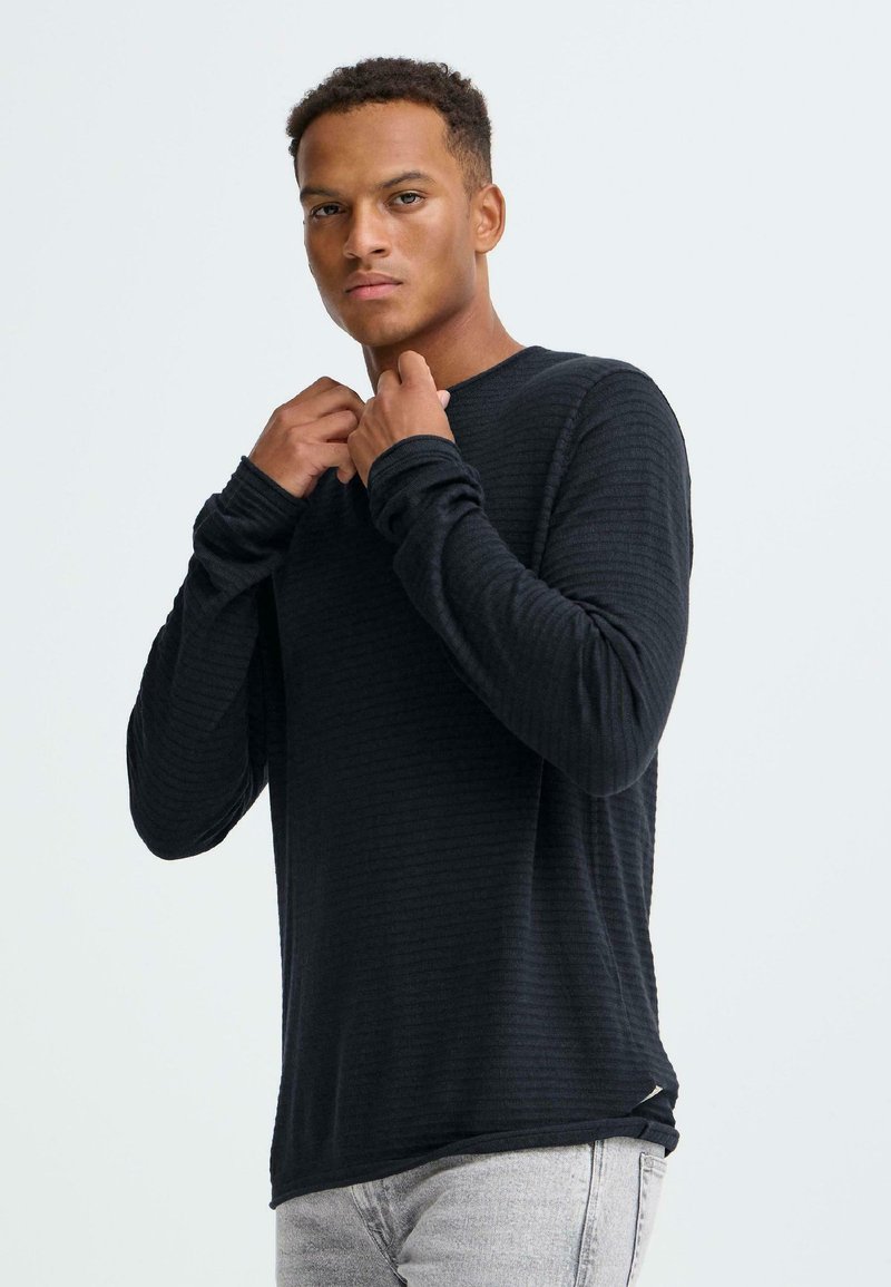 Zwart shirt met lange mouwen en ribbelstructuur, ronde halslijn en een slanke pasvorm, gecombineerd met lichtgrijze spijkerbroek.