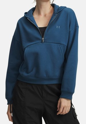 Femme portant un sweat à capuche bleu avec fermeture éclair partielle, poche avant et logo Under Armour, associé à un pantalon noir.