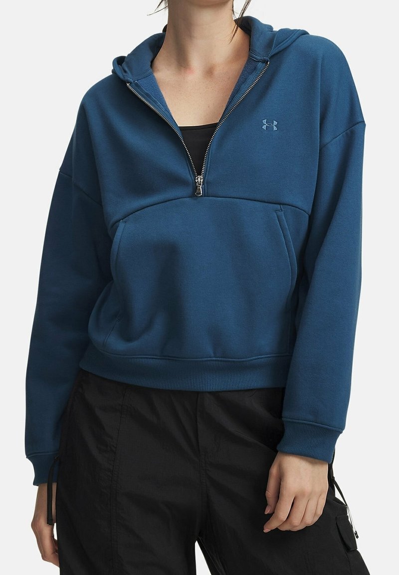 Femme portant un sweat à capuche bleu avec fermeture éclair partielle, poche avant et logo Under Armour, associé à un pantalon noir.