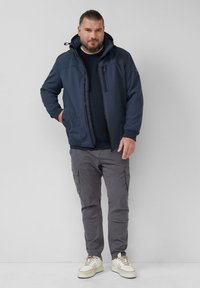 Donkerblauwe hoodie met een rits aan de voorkant, zijzakken en een zwart pocketdetail. Draag het over een gebreide trui en grijze cargo pants, met sneakers.