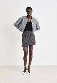 Chaqueta bomber gris con botones de presión en el frente, blusa negra con encaje, minifalda a rayas y zapatos de tacón blanco.