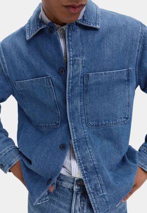 Denimjakke i medium blå med knaplukning foran, spids krave og to store brystlommer. Tekstureret stof med synlige syningsdetaljer.
