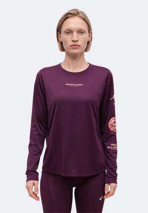 Γυναίκα που φορά μακρυμάνικη μωβ μπλούζα ASICS FUJITRAIL με λογότυπα στο στήθος και το μανίκι, στέκεται μπροστά σε ένα απλό φωτεινό φόντο.