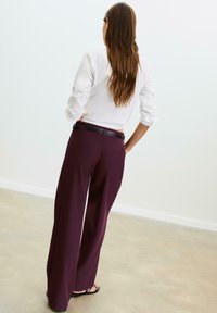 Pantaloni a gamba larga in un profondo bordeaux, con una superficie liscia, una vita in pelle e due tasche posteriori. Il modello indossa una maglietta aderente bianca.
