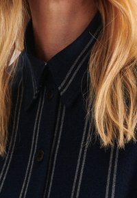 Marinblå kragskjorta med vita vertikala ränder, med knappstängning fram och en mjuk struktur. Kragen ligger platt mot nacken.