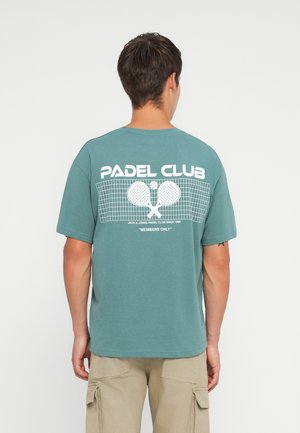 Teal cotton T-shirt with "PADEL CLUB" printed in white on the back, featuring a graphic of rackets and a net. Short sleeves, round neckline.