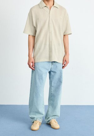 Light beige short-sleeve shirt with a textured pattern and collar, paired with loose light blue denim jeans and beige sneakers.