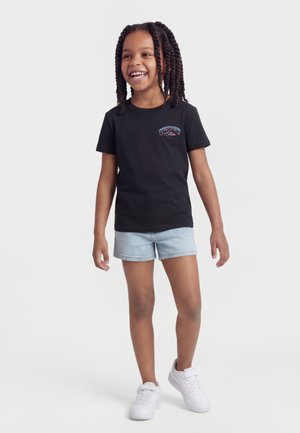 Ragazza giovane con capelli intrecciati che sorride, indossa una t-shirt nera Quiksilver, shorts in denim chiaro e scarpe da ginnastica bianche, in piedi su uno sfondo bianco.