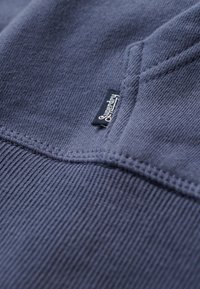 Mørkeblå stof med struktureret ribbehæftning. Har et lille sort mærke med "Superdry" i hvidt på sømmen. Nærbillede af syningsdetalje.