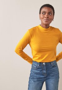 Langærmet, tætsiddende top i lys gul bomuld med høj hals, parret med højtaljede blå denimjeans med lys syning.