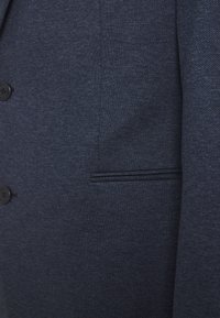 Blazer blu navy con una trama testurizzata, dotato di una tasca sul petto e tre bottoni neri sulla parte anteriore.