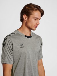 Jeune homme aux cheveux bruns courts portant un maillot de sport gris Hummel avec des logos chevrons noirs sur les épaules, regardant vers la droite sur un fond uni.