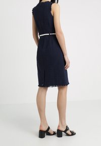 Námořnická modrá bezramínková šaty s texturovanou tkaninou, třásněmi na výstřihu a lemu a bílým opaskem jako akcentem. Nošeno s černými sandály na širokém podpatku.