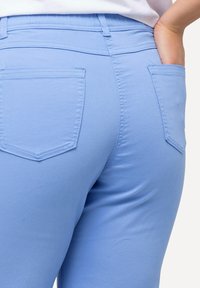 Vaaleansiniset joustavat housut, jotka istuvat tiukasti ja joissa on kaksi takataskua sekä näkyviä ompelutaitoksia. Tasainen kankaan pinta.