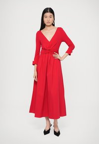 Červené zavinovací šaty s dlouhým rukávem, hlubokým výstřihem do V, zvýrazněným pasem a splývavou sukní. Vyrobené z měkké látky; modelka má na sobě černé špičaté podpatky.