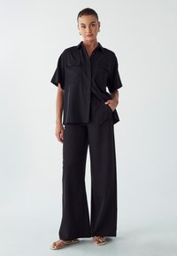 Černý dvoudílný outfit, který zahrnuje košili s krátkým rukávem na knoflíky se dvěma náprsními kapsami a kalhoty širokého střihu, vyrobené z hladkého materiálu.
