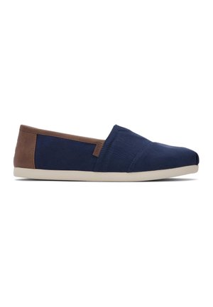 Zapato casual tipo slip-on con parte superior de tela azul marino, panel trasero marrón y suela beige, mostrado de lado sobre fondo blanco.