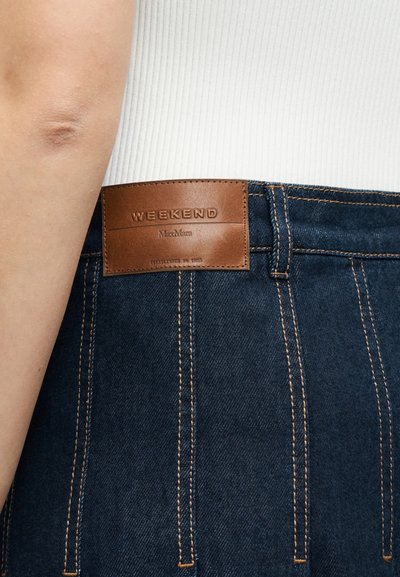 Δερμάτινη ετικέτα μάρκας με την επιγραφή "WEEKEND MaxMara" σε σκούρο μπλε τζιν παντελόνι, φορεμένο με λευκό ριμπ τοπ.