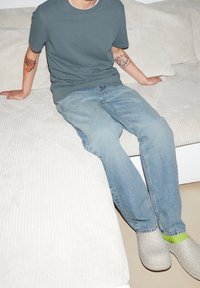 Šedé tričko, světle modré džíny a šedé pletené slip-on boty se zelenými akcenty. Sedí na strukturované bílé pohovce s polštáři.