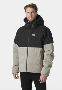 Gevoerde jas met een zwart bovenstuk en een beige onderstuk, voorzien van een capuchon, ritssluiting aan de voorkant en een gestructureerde stof voor isolatie.