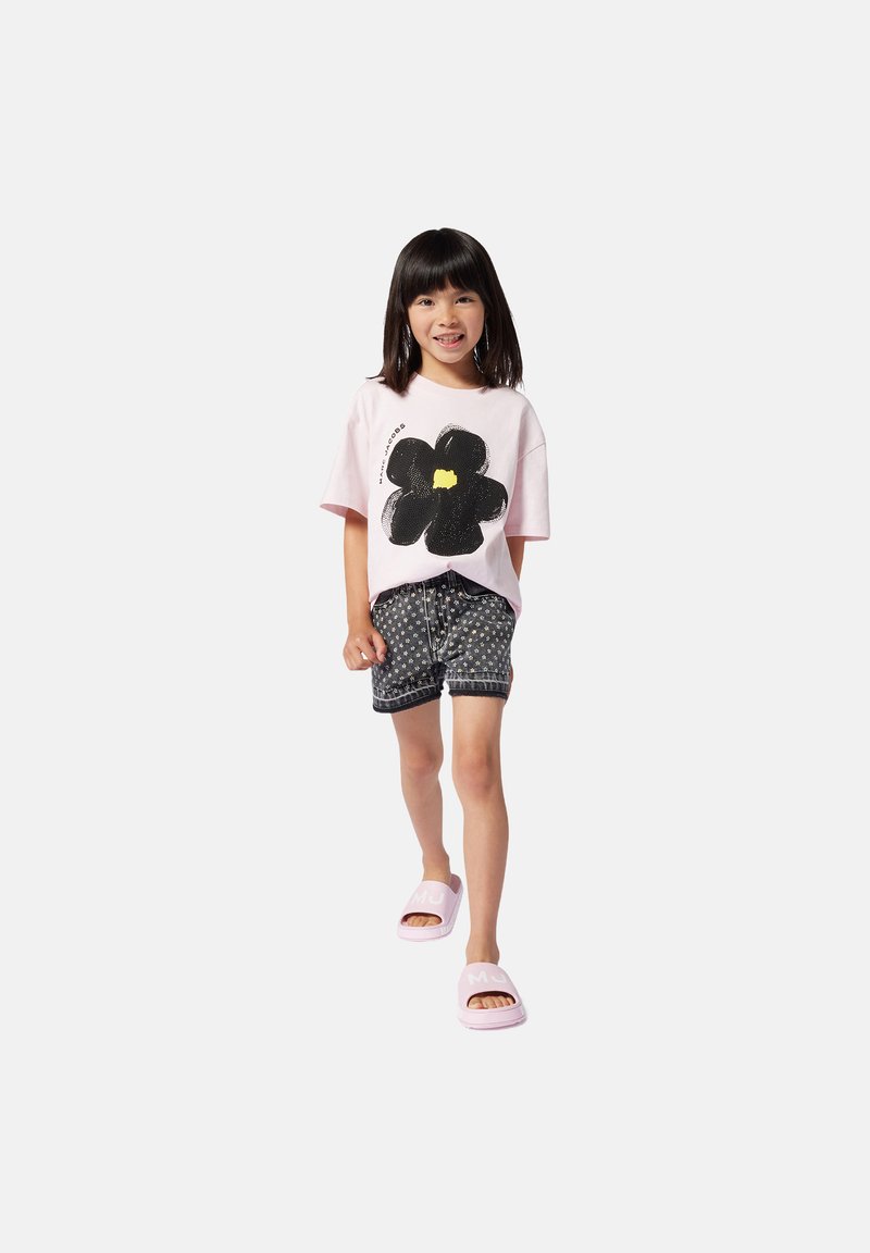 T-shirt rosa chiaro con una grafica di fiore nera, abbinata a pantaloni corti scuri con fantasia. Indossato con sandali scivolosi rosa. Outfit semplice e casual.