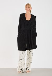 Rochie de baie neagră, pufoasă, cu glugă, curea cu legare proprie și buzunare laterale. Purtați deasupra unei pijamale albe cu pantaloni ce prezintă modele celestiale negre.