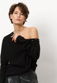 Schwarzer, schulterfreier Pullover mit langen Ärmeln, der silberfarbene Akzente entlang des Ausschnitts und einen weichen, strukturierten Stoff aufweist.