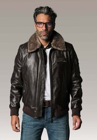Veste bomber en cuir marron avec un col en fourrure, des poignets et une taille côtelés. Comprend une poche poitrine et une poche zippée sur le bras. Portée par-dessus une chemise blanche.
