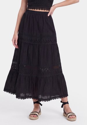 Woman wearing a black eyelet lace midi skirt with scalloped edges and black ankle-tie espadrille sandals against a white background.