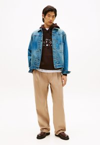 Τζιν μπουφάν συνδυασμένο με καφέ φούτερ με λευκή επωνυμία, ανοιχτόχρωμο παντελόνι και σκούρα καφέ παπούτσια. Απλό λευκό φόντο.