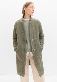 Olivgrüner Boucle-Mantel mit einer vorderen Knopföffnung, Seitentaschen und einem lässigen Schnitt, kombiniert mit einem hellgrünen strukturierten Pullover.