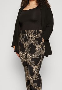 Svart kardiagan över en svart topp, i kombination med svarta byxor med en beige bladtryck. Tyget verkar mjukt med en slät yta.