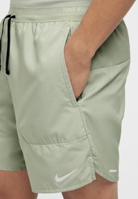 Calções de corrida Nike verde-claro com cordão preto, painel lateral em rede e logotipo da Nike branco na bainha. Textura suave e ajuste atlético.