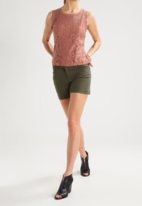 Top sin mangas de encaje rosa con un patrón texturizado, combinado con pantalones cortos verdes oliva y botas de tacón abiertas en la parte delantera de color negro con cremalleras.