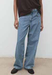 Vaqueros de pierna ancha azul claro con un acabado texturizado, que presentan una cintura alta y un diseño clásico de cinco bolsillos. Combinados con una blusa marrón.