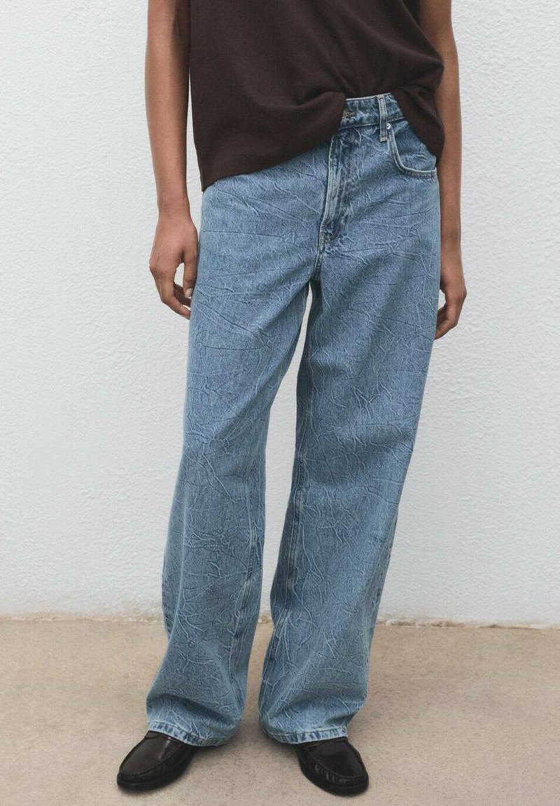 Vaqueros de pierna ancha azul claro con un acabado texturizado, que presentan una cintura alta y un diseño clásico de cinco bolsillos. Combinados con una blusa marrón.