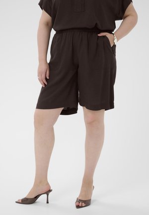 Personne portant un short noir ample et un haut assorti, posant avec une main dans la poche et portant des sandales à talons hauts marron.