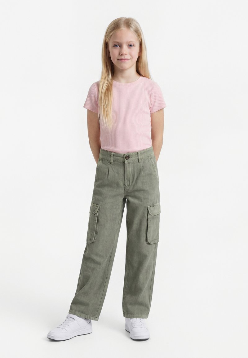 Jeune fille aux longs cheveux blonds debout, portant un t-shirt côtelé rose clair, un pantalon cargo vert olive et des baskets blanches sur fond blanc.