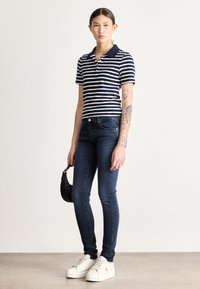 Pruhované polo tričko v námorníckej modrej a bielej farbe, priliehavý strih, v kombinácii s tmavomodrými úzkymi džínsami a bielymi teniskami, držiaca malú čiernu kabelku.