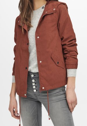 Femme portant une veste à capuche couleur rouille sur un pull gris clair et un jean gris taille haute avec plusieurs boutons visibles.