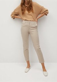 Pull beige en maille côtelée, pantalon beige fuselé et chaussures blanches à enfiler, sur un fond blanc uni. Le pantalon a une texture lisse.