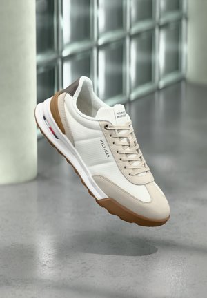 Sneaker Tommy Hilfiger bianca e beige con suola marrone, logo marchiato sul lato e sulla linguetta, sospesa sopra un pavimento grigio liscio.
