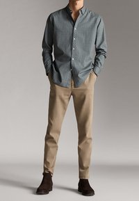 Camicia button-down a quadretti blu e bianchi, abbinata a chinos beige e stivali alla caviglia marrone scuro. Tessuto morbido, vestibilità su misura.