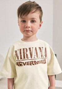 T-shirt à manches courtes couleur crème, avec un grand texte rétro marron indiquant "NIRVANA" et "NEVERMIND", offrant une coupe décontractée.