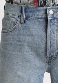 Vaqueros de denim azul claro con un diseño clásico de cinco bolsillos, que presentan costuras visibles, herrajes de botón de latón y una textura ligeramente desgastada.