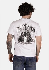 T-shirt bianco in cotone con una grande grafica di un serpente nero sul retro, con dettagliati squame e la parola "PROPAGANDA" in carattere grassetto sopra.