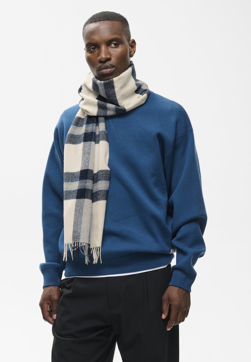 Homme portant un sweat-shirt bleu et un pantalon noir avec une écharpe à carreaux beige, noire et grise enroulée autour du cou sur un fond uni.