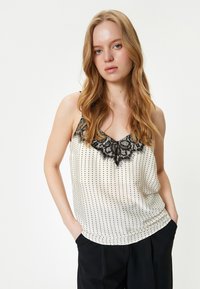 Top sin mangas color crema con lunares negros, con un borde de encaje en el escote. Textura suave; corte relajado. Combinado con pantalones negros.
