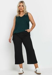 Top senza maniche verde con scollatura a V, abbinato a pantaloni neri a gamba larga e sneakers grige con accenti neri. Outfit semplice e informale.